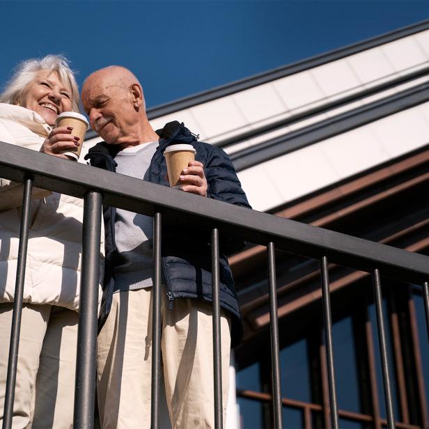 Ein älteres Paar steht lächelnd mit Kaffeebechern auf dem Balkon eines modernen Hauses.