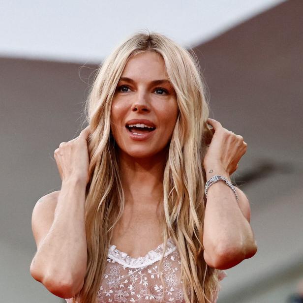 Sienna Miller mit blonden, gewellten Haaren und durchsichtigem Oberteil lächelt.