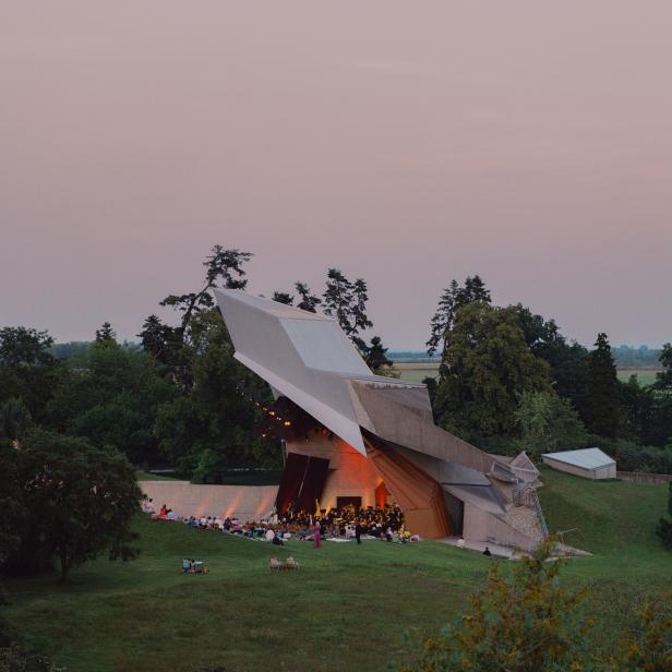 Eine moderne Freilichtbühne mit ungewöhnlicher Dachkonstruktion steht inmitten einer grünen Parklandschaft.