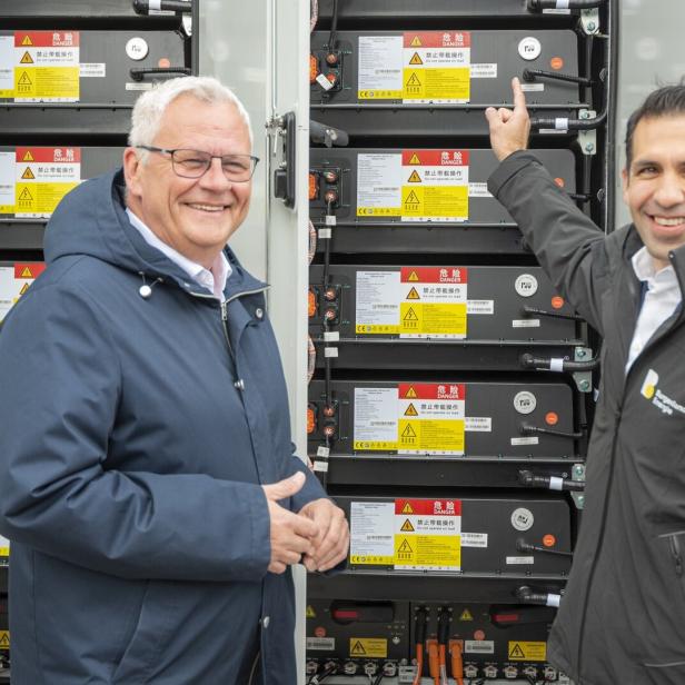 Zwei Männer stehen vor mehreren großen Batteriespeichern mit Warnhinweisen und lächeln, einer zeigt auf eine Batterie.