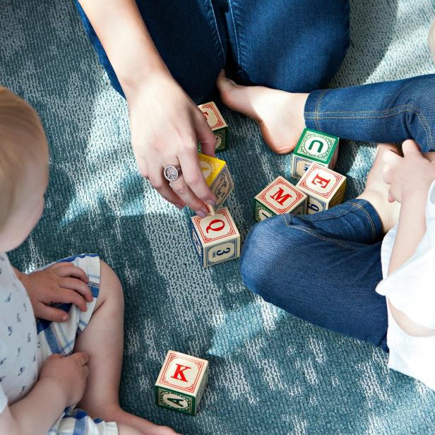 Zwei kleine Kinder und eine erwachsene Person spielen gemeinsam mit bunten Holzbausteinen auf dem Teppich.