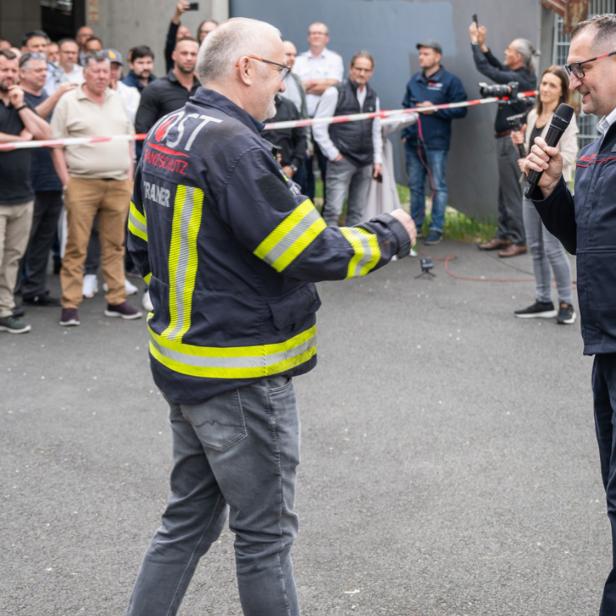 Zwei Männer in Feuerwehrkleidung sprechen vor einer größeren Menschenmenge im Freien, einer hält ein Mikrofon.