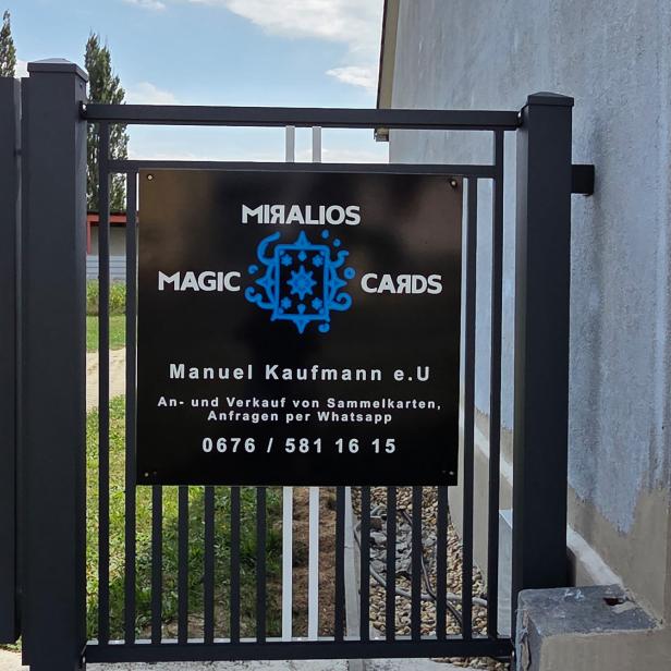 Schwarzes Firmenschild von „Miralios Magic Cards“ an einem modernen Metalltor vor einem Wohnhau