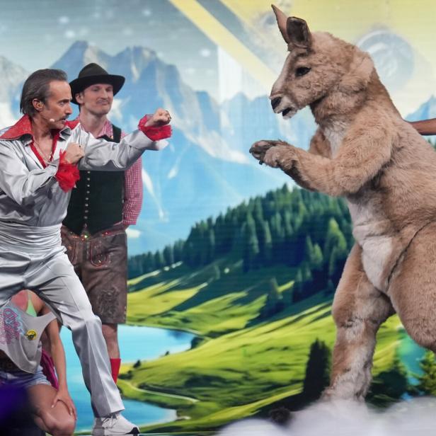 Mehrere Menschen in bunten Kostümen stehen vor einer Alpenlandschaft, einer davon kämpft spielerisch mit einem Känguru im Kostüm.