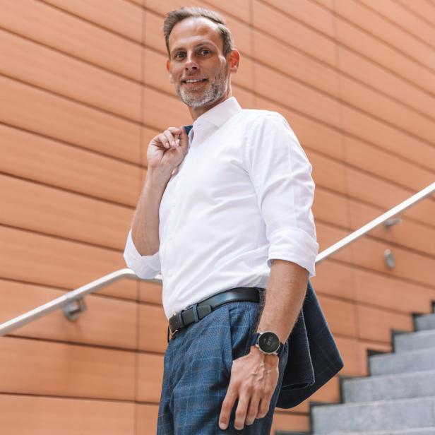 Ein Mann in Businesskleidung steht auf einer Treppe vor einer modernen Wandverkleidung und lächelt.
