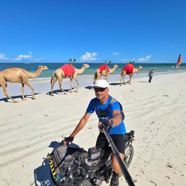 Ein Mann macht ein Selfie auf einem Fahrrad am Strand, während mehrere Kamele mit roten Decken am Meer entlanggeführt werden.