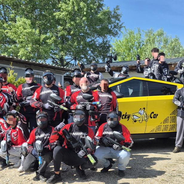 Gruppe von Paintballspielern in Ausrüstung vor einem gelben Auto im Freien bei sonnigem Wetter.