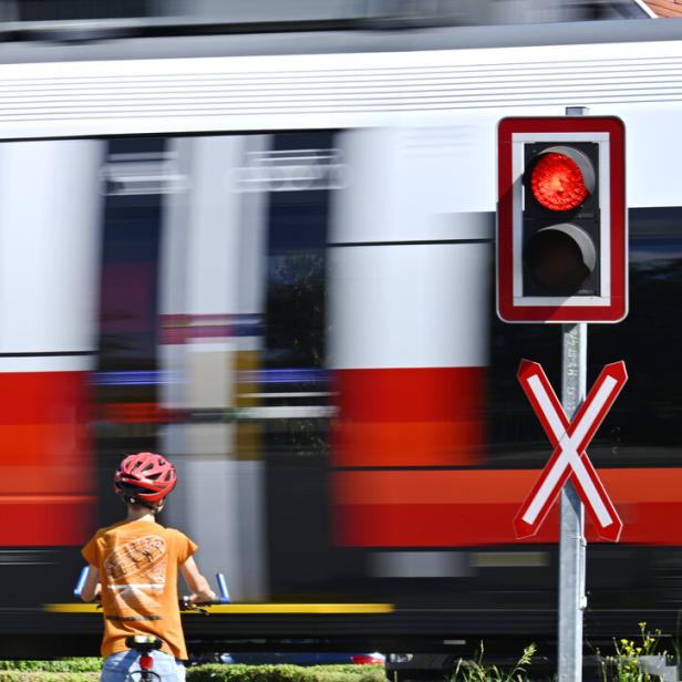 Ein Kind mit Fahrrad wartet an einem Bahnübergang mit rotem Signal, während ein Zug vorbeifährt.