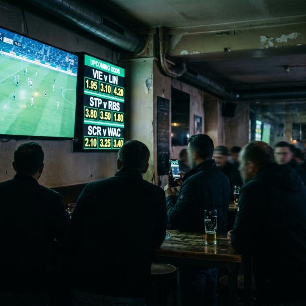 Dunkle Bar mit mehreren Personen, die auf eine große Wandbildschirm mit Fußballspiel und daneben angezeigten Wettquoten schauen