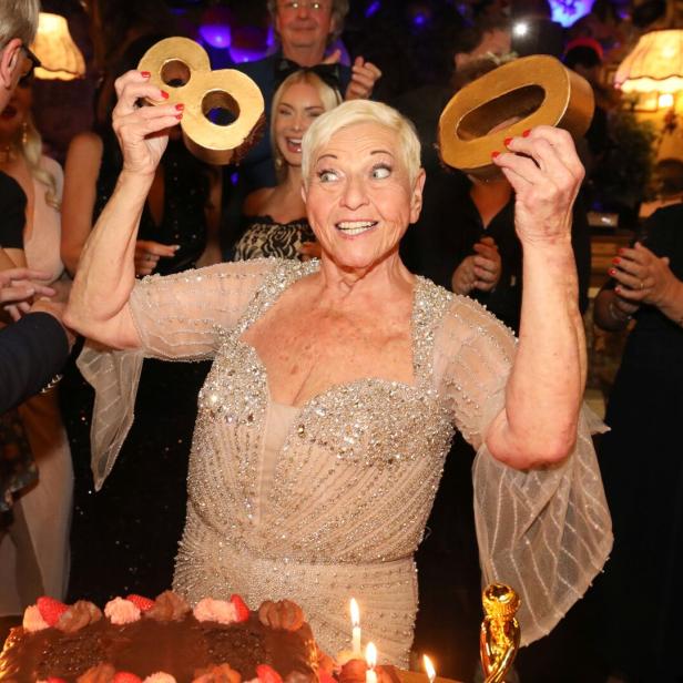 Eine strahlende Frau hält goldene Zahlen über einem Geburtstagskuchen mit Kerzen.