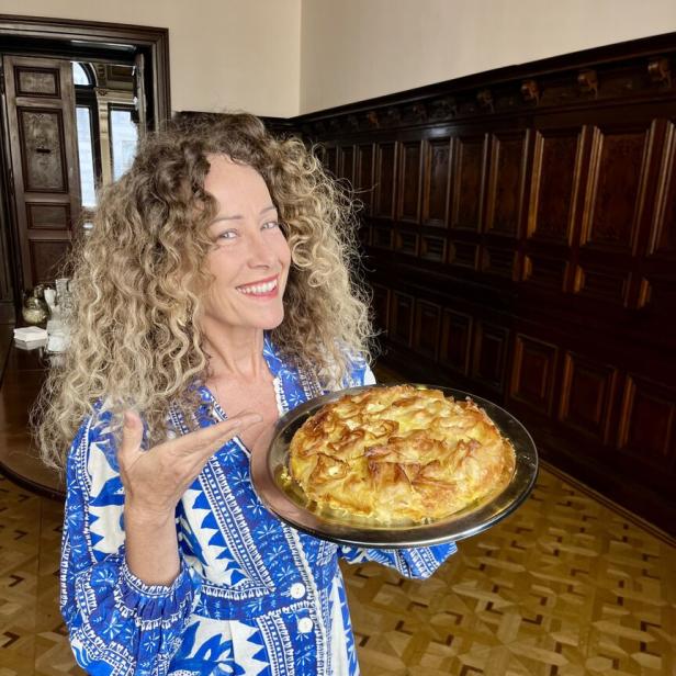 Eine Frau mit lockigem Haar präsentiert lächelnd einen runden Kuchen auf einem silbernen Tablett.