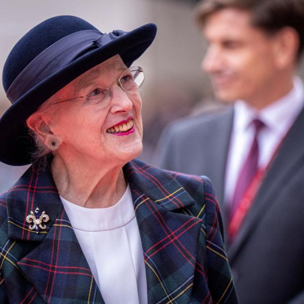 Königin Margrethe II. lächelt in einem karierten Blazer und Hut, während ein Mann im Anzug im Hintergrund steht.