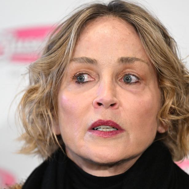 Sharon Stone mit blonden, gewellten Haaren und dunklem Rollkragenpullover blickt aufmerksam zur Seite.