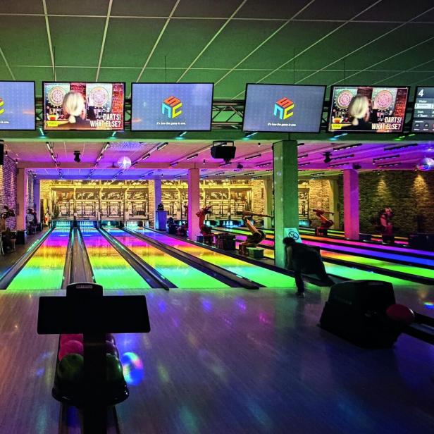 Mehrere Bowlingbahnen mit bunten Lichtern, Menschen beim Bowling und Bildschirmen über den Bahnen.