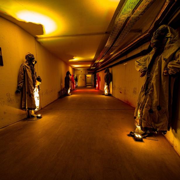 Ein langer, gelb beleuchteter Gang mit Schaufensterpuppen in Schutzanzügen und Gasmasken an den Wänden.