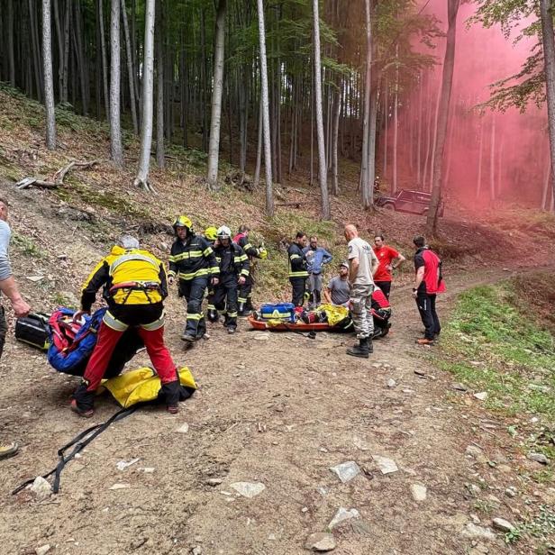Mehrere Rettungskräfte und Helfer versorgen eine Person auf einer Trage im Wald, während im Hintergrund roter Rauch aufsteigt.