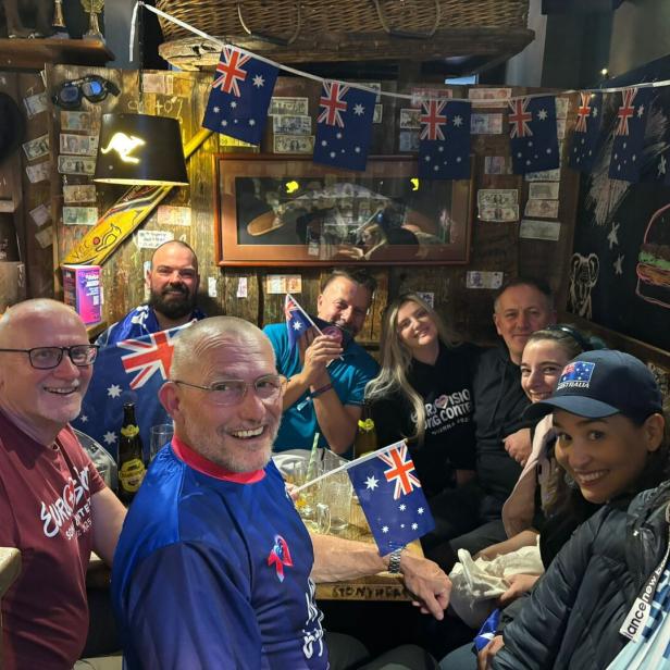 Eine fröhliche Gruppe von Menschen sitzt in einem dekorierten Raum mit vielen australischen Flaggen und lächelt in die Kamera.