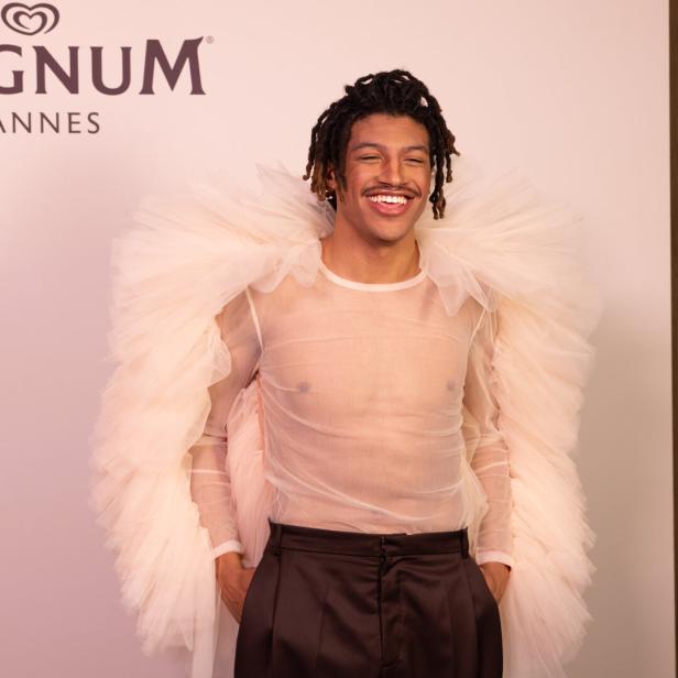 Ein lächelnder Mann mit Dreadlocks trägt ein transparentes Oberteil und einen bauschigen Kragen vor einem Magnum Cannes Logo.