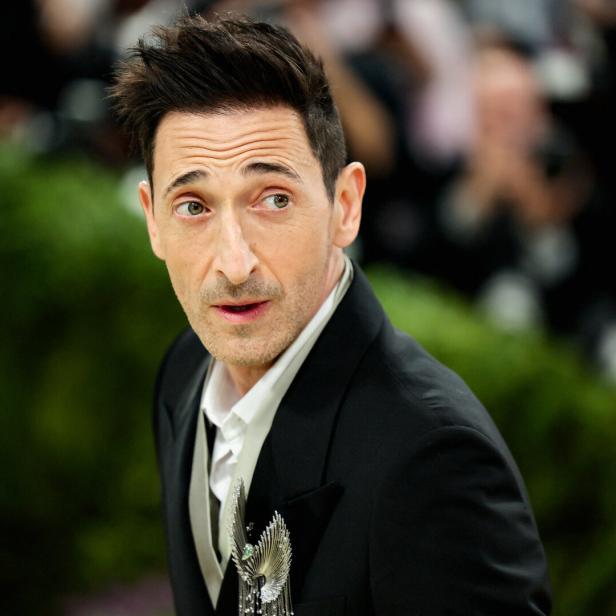 Adrien Brody im schwarzen Anzug mit Brosche blickt aufmerksam zur Seite.