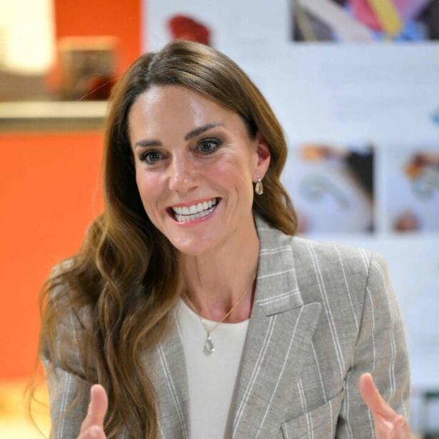 Kate, mit Blazer, lächelt und gestikuliert mit beiden Händen.