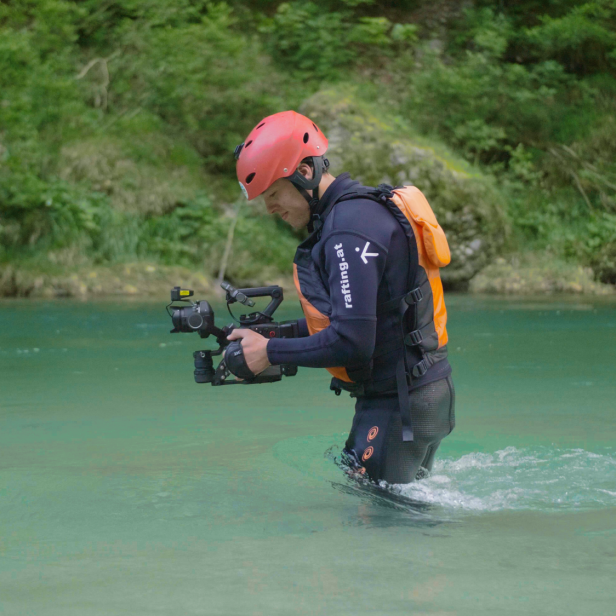 Eine Person mit Helm und Kamera steht im flachen Wasser eines türkisfarbenen Flusses
