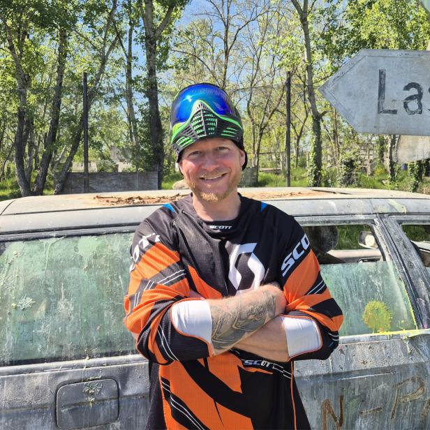 Mann in Paintball-Ausrüstung steht vor einem alten Auto und einem Wegweiser nach Lassee.