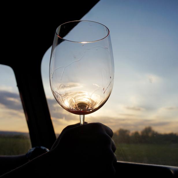 Eine Person hält bei Sonnenuntergang ein leeres Weinglas vor ein Fahrzeugfenster.