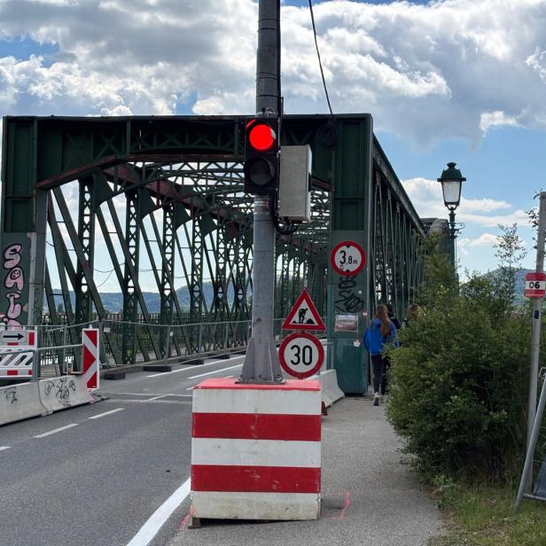 Eine grüne Stahlbrücke mit Baustellenabsperrungen, Verkehrsschildern und roter Ampel, zwei Personen gehen auf dem Gehweg.