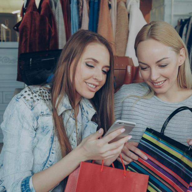 Zwei Frauen sitzen nebeneinander, eine hält ein Smartphone, die andere eine gestreifte Einkaufstasche