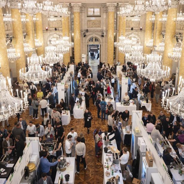 Viele Menschen besuchen eine Messe mit Ständen in einem prunkvollen Saal mit großen Kronleuchtern und goldenen Säulen.