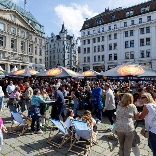 Viele Menschen sitzen und stehen an Tischen unter Sonnenschirmen auf einem belebten Platz inmitten historischer Gebäude.