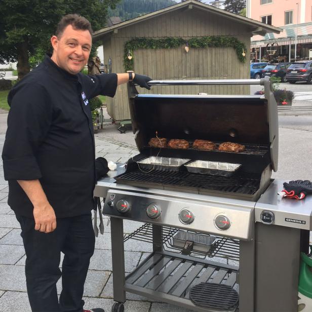 Ein Mann grillt Fleisch auf einem Gasgrill im Freien vor einem Holzhaus und geparkten Autos.