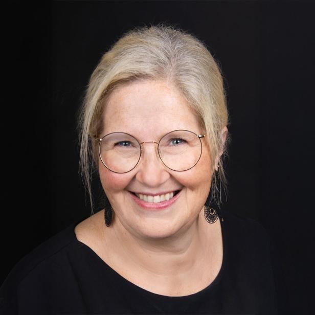 Frau mit blondem Haar, Brille und schwarzem Oberteil lächelt vor schwarzem Hintergrund.