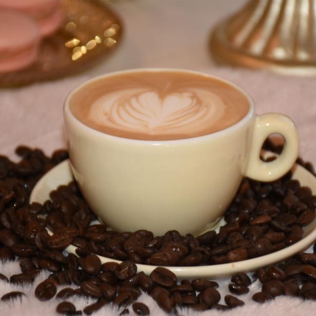 Cappuccino mit Latte Art in einer Tasse, umgeben von Kaffeebohnen und rosa Macarons im Hintergrund.