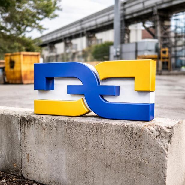 Blau-gelbes DDZ-Logo auf Betonmauer vor Industrie- und Baustellenkulisse.