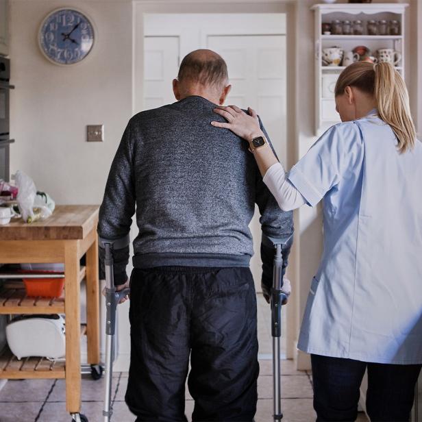 Pflegekraft unterstützt älteren Mann mit Gehhilfen in einer hellen Küche.