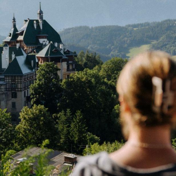 Blick über die Schulter einer Frau mit zusammengebundenem Haar auf ein Schloss in einer bewaldeten Hügellandschaft.