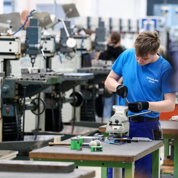 Ein junger Mann arbeitet konzentriert an einem Schraubstock in einer Werkstatt mit mehreren Maschinen im Hintergrund.