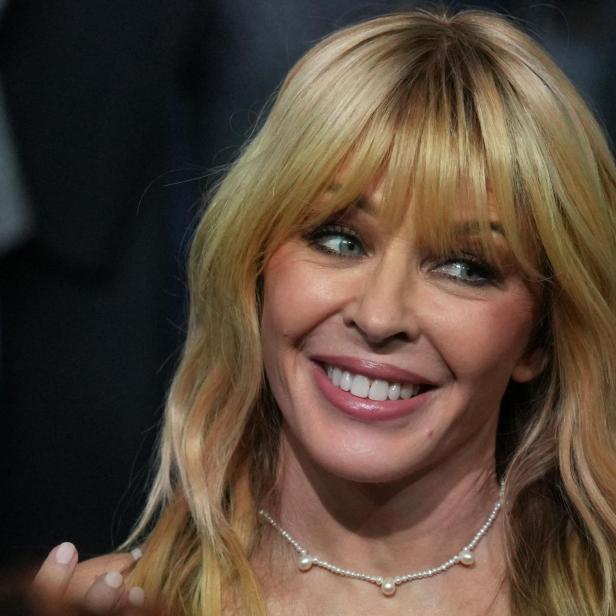 Kylie Minogue mit blondem Haar und Pony lächelt, trägt eine Perlenkette und steht vor unscharfem Hintergrund.