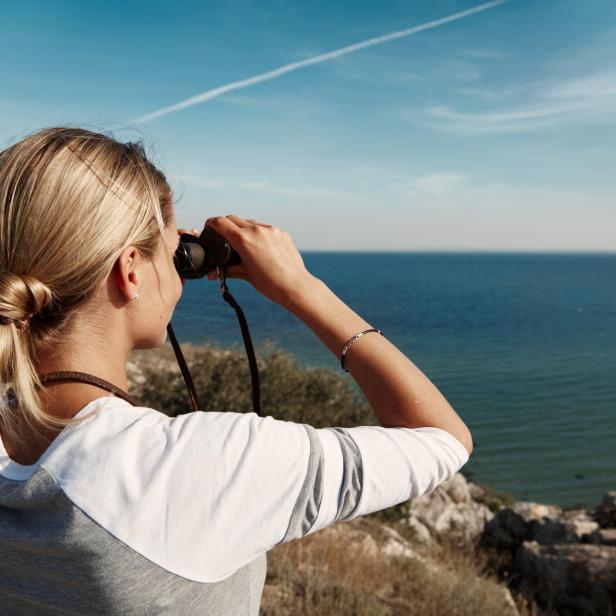 Frau mit Fernglas blickt von einer Küste aus über das Meer in die Ferne.