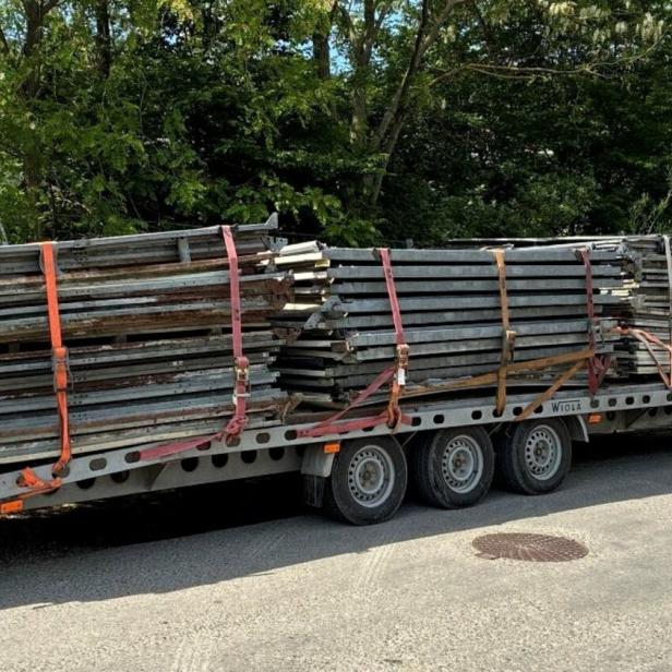 Ein weißer Transporter zieht einen Anhänger mit gestapelten Metallteilen, die mit Gurten gesichert sind.