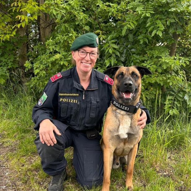 Eine Polizistin in Uniform kniet neben einem Polizeihund, beide lächeln und sitzen vor grünem Gebüsch.