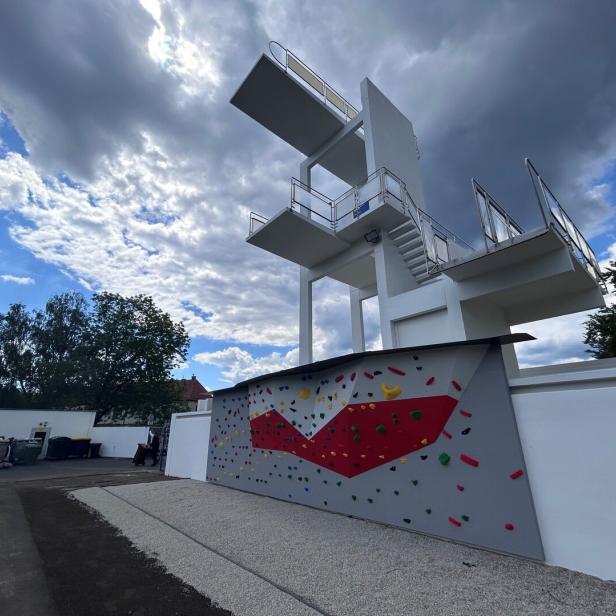 Ein hoher Sprungturm mit mehreren Plattformen steht neben einer bunten Kletterwand im Freien unter bewölktem Himmel.