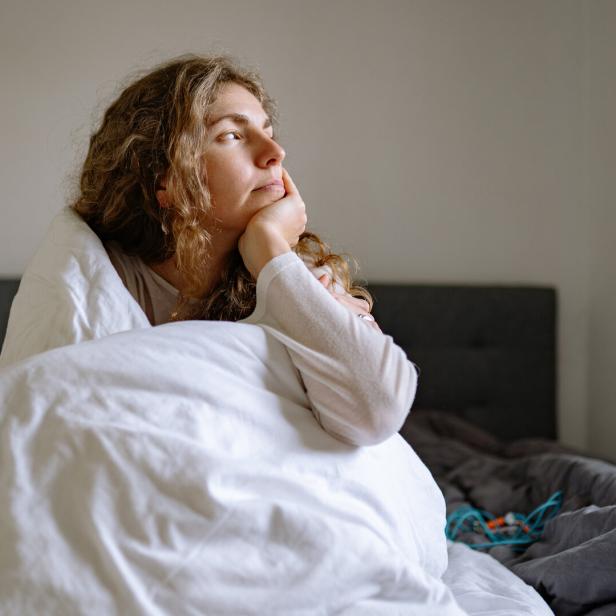 Eine Frau liegt nachdenklich unter einer weißen Decke im Bett und blickt aus dem Fenster.
