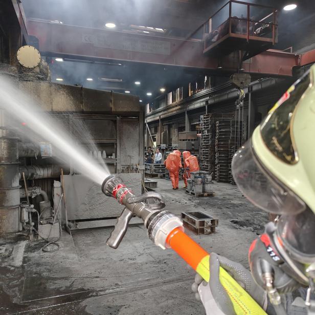 Ein Feuerwehrmann spritzt Wasser in einer Industriehalle, im Hintergrund arbeiten Personen in orangefarbenen Schutzanzügen.