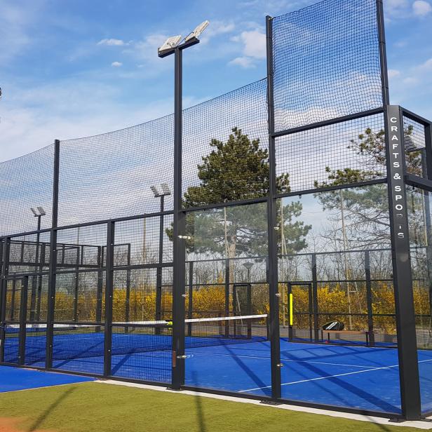 Ein moderner Padelplatz im Freien mit blauen Spielfeldern und hohen Glas- und Gitterwänden unter blauem Himmel.