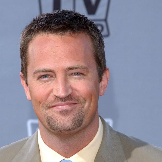 Matthew Perry lächelt mit kurzem, dunklem Haar und leichtem Bart vor grauem Hintergrund.