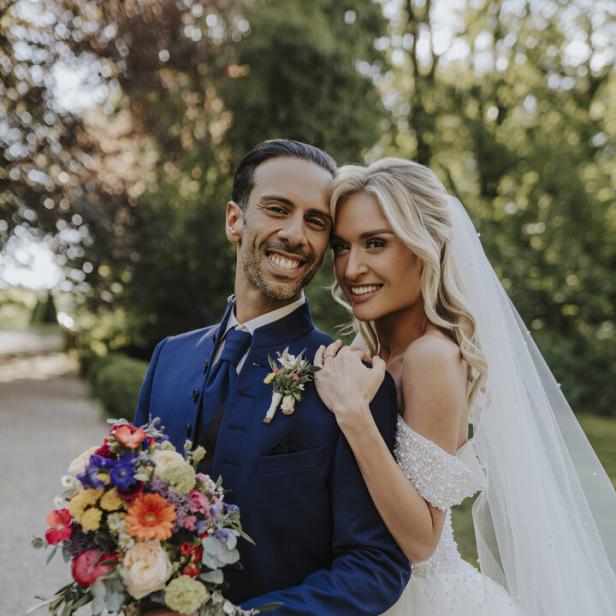 Ein lächelndes Brautpaar steht eng umarmt im Freien, die Braut trägt ein weißes Kleid und der Bräutigam hält einen bunten Blumenstrauß.