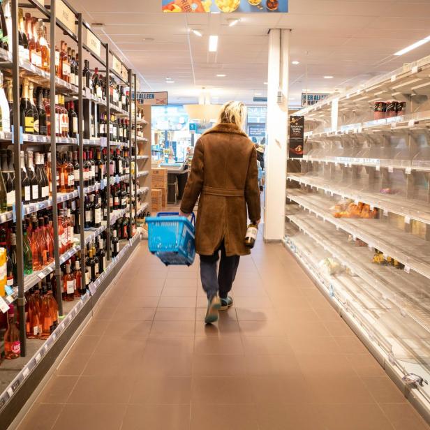 Person mit Einkaufskorb geht durch einen Supermarktgang mit leeren und vollen Regalen.