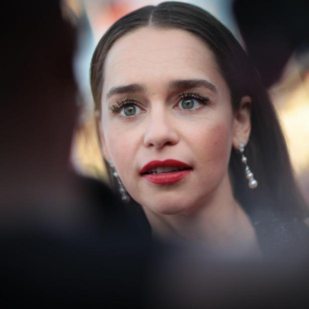 Emilia Clarke mit roten Lippen und Ohrringen schaut aufmerksam zur Seite.